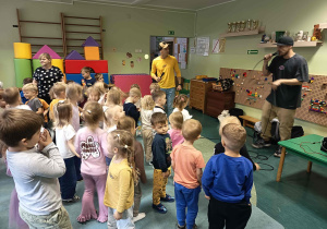 Przedszkolaki stoją zapatrzone w muzyków prowadzących koncert.