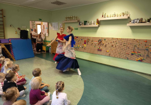 Tancerki w stylizowanych strojach, które tańczą przed grupą dzieci siedzących na dywanie.