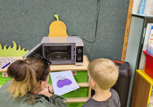 Chłopiec razem z panią nauczycielką wkłada pracę do mikrofalówki.