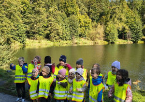 Przedszkolaki na tle stawu machają śpiewając jesienne piosenki.