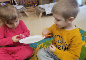 Chłopiec z dziewczynką podają sobie talerzyk papierowy za pomocą spinacza do bielizny.