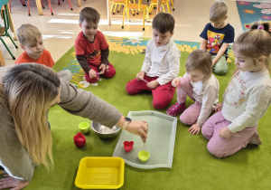 Nauczycielka pokazuje dzieciom jak napełnić wodą foremki za pomocą pipetki.