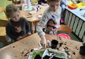 Chłopiec za pomocą łyżki sypie ziemię do doniczki.