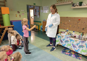 Warsztaty chemiczne dla dzieci w przedszkolu, podczas których prowadząca pokazuje przedszkolakom bezpieczne eksperymenty.